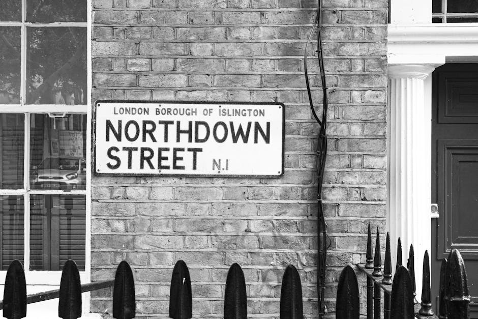 A black and white image showing two white box trucks parked on a curved cobblestone street in an urban area. The trucks are positioned parallel to a brick wall on the right side, with their rear doors