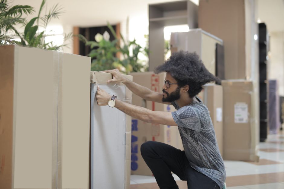 Two individuals are engaged in packing for an office relocation within an indoor space. The person on the left, wearing a white shirt, is seated on the floor, handling a cardboard box labeled 'OFFICE'