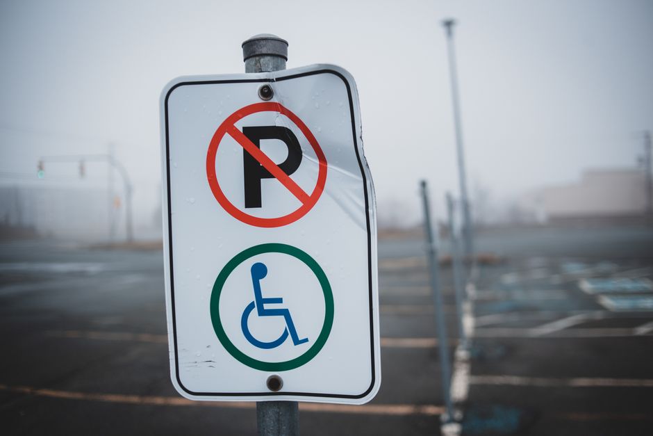 Close-up photograph of a parking sign affixed to a metal pole in an outdoor parking area, with a blurred backdrop of empty parking spaces and concrete pavement. The sign displays a red circle with a d