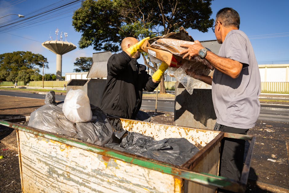 Two men are engaged in a waste disposal activity at an outdoor recycling or waste collection point, with a large, portable metal container filled with black plastic garbage bags and cardboard, some of